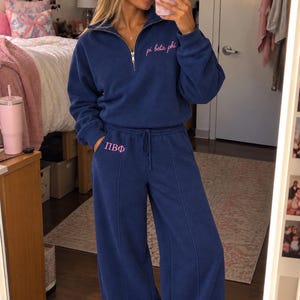Peut inclure: Un ensemble assorti bleu marine composé d'un pull à quart de zip et d'un pantalon de jogging large. Le pull présente le texte pi beta phi brodé en rose sur la poitrine. Le pantalon présente les lettres grecques ΠΒΦ brodées en rose sur la hanche.
