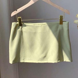 Pu&ograve; includere: Una minigonna color verde lime pallido con orlo dritto e superficie liscia priva di decorazioni. La gonna &egrave; appesa a una gruccia in legno chiaro tramite due clip metalliche dorate, presentando un design semplice e strutturato senza tasche.