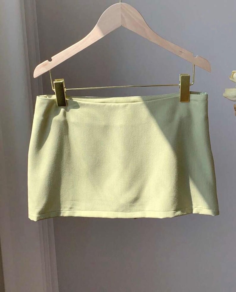 Pu&ograve; includere: Una minigonna color verde lime pallido con orlo dritto e superficie liscia priva di decorazioni. La gonna &egrave; appesa a una gruccia in legno chiaro tramite due clip metalliche dorate, presentando un design semplice e strutturato senza tasche.