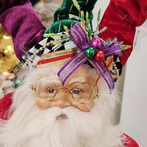 May include: A whimsical Christmas figurine of a Santa Claus with a white beard and glasses. He is wearing a red and green velvet hat with a purple ribbon and gold bells. The hat is decorated with green sprigs and a black and white checkered pattern.