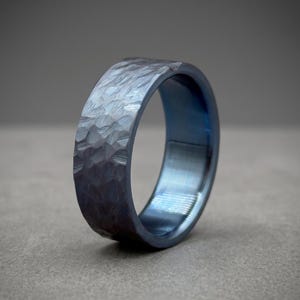 May include: A dark blue metal ring with a wide band and a textured, hammered finish on the exterior surface. The interior of the band features a smooth, polished finish with a deep blue metallic sheen, creating a contrast with the outer texture.