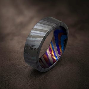May include: A ring featuring a dark grey, layered exterior with a visible wood-grain-like pattern. The interior of the band displays a vibrant, multi-coloured finish with distinct wavy stripes in shades of blue, purple, and copper-orange.