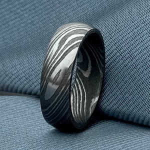 Pode incluir: Um anel de metal prateado e preto com um padrão. O anel é largo e tem um acabamento liso e polido.