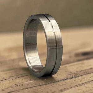 May include: A brushed silver ring with a centered, thin, dark groove. The ring has a smooth, matte finish and a simple, modern design. The ring is displayed on a wooden surface.