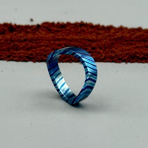 May include: A blue and white striped ring with a unique, open design. The ring is made of a material that appears to be resin or a similar composite. The ring is set against a neutral gray background with a blurred brown texture in the background.