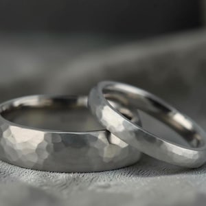 May include: Two silver rings with a hammered texture. One ring is wider with a rounded band, and the other is thinner with a smooth band. The rings are displayed on a gray surface.
