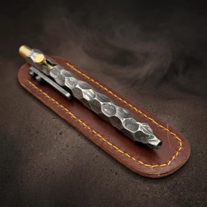 May include: A Damascus steel bolt-action pen with a hammered, textured surface, a gold-colored metal bolt mechanism, and a matching metal pocket clip, resting on a brown leather sleeve with yellow stitching along the rounded edges.