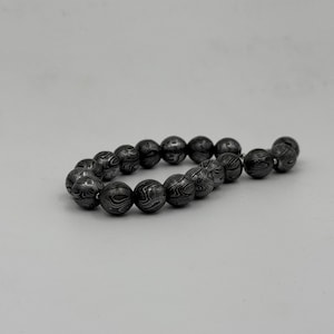 May include: A dark gray beaded bracelet. The beads are round with a patterned surface. The bracelet is made of a dark gray material and is displayed against a white background. The beads have a unique, swirling design.
