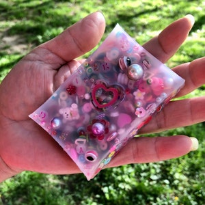 Op de afbeelding: Een heldere roze harsblok gevuld met kleurrijke kralen, bedels en een roze hart. Het blok wordt in een hand gehouden.