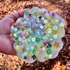 Puede incluir: Un posavasos de resina transparente con una variedad de cuentas de colores y verdes incrustadas en su interior. El posavasos tiene forma de nube y un acabado liso y brillante.