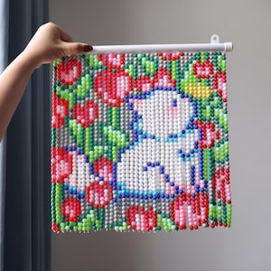 May include: A square wall hanging composed of small, colorful plastic beads arranged to form a pixelated image of a white cat surrounded by red, pink, and green floral patterns. The bead art is suspended from a white plastic rod with a hanging loop.