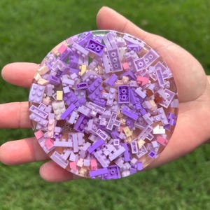 May include: A circular coaster made of clear resin embedded with a dense collection of small plastic building bricks in various shades of purple, lavender, and light pink. The surface is smooth and glossy, showcasing the layered, multi-dimensional arrangement of the interlocking brick pieces throughout the transparent material.