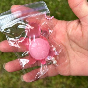 May include: A small, translucent pink turtle-shaped object encased in a clear plastic zip-top bag. The turtle features a smooth, rounded shell and four flippers. A short string of small, iridescent white beads is attached to the turtle figure.