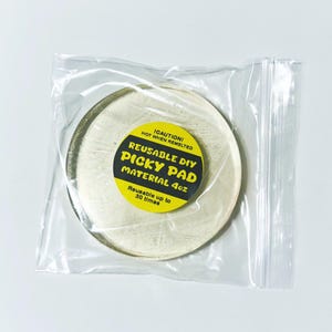 Op de afbeelding: Een ronde, crèmekleurige, herbruikbare DIY Picky Pad, 4oz, in een doorzichtige plastic zak. De pad heeft een geel en zwart label met de tekst "REUSABLE DIY PICKY PAD MATERIAL 4oz" en "Reusable up to 30 times".