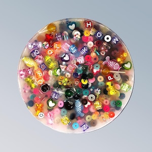 Könnte beinhalten: Ein runder, transparenter Harz-Untersetzer mit einer Vielzahl von bunten Perlen, Buchstaben und Herzen, die darin eingebettet sind. Der Untersetzer ist in einer Mischung aus Farben gehalten, darunter Rot, Blau, Grün, Gelb, Rosa und Lila.