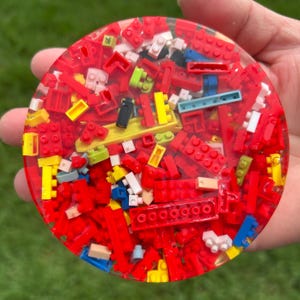 May include: A circular coaster made of clear resin embedded with a dense collection of small plastic building bricks in various colors, primarily red, with accents of yellow, white, blue, green, and black pieces visible throughout the surface.
