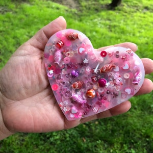 Puede incluir: Una pieza de resina en forma de corazón rosa llena de cuentas de colores, dijes y pequeños peces de plástico. La resina es transparente y los dijes son visibles.