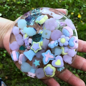 May include: A clear, circular resin piece filled with pastel-coloured charms. The charms include stars, flowers, hearts, and other shapes in shades of blue, yellow, pink, and purple. Some charms have the word "Sweet" on them.