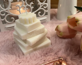 May include: A white candle shaped like a stack of square books with a small gear-like topper, a white metal lantern, a white ceramic pitcher, two pale pink tulips, and a copper-colored metal candle snuffer with a long handle.