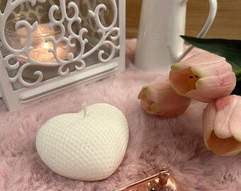 May include: A white heart-shaped candle with a textured grid pattern sits on a soft pink surface. Nearby are a white decorative lantern, a small white ceramic pitcher, two pale pink tulip flowers, and a rose gold metal candle wick snuffer.