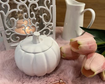 May include: A white ceramic pumpkin-shaped lidded jar, a white metal lantern with a lit candle, a white ceramic pitcher, a copper-colored metal candle snuffer, and three pale pink artificial tulips arranged together in a decorative display.