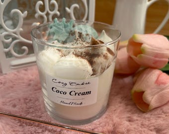 May include: A clear glass candle container filled with white wax, topped with white meringue-like wax decorations and a dusting of brown powder. A white label on the front reads Cozy Ember Coco Cream Hand Made. A copper-colored metal candle snuffer is present.