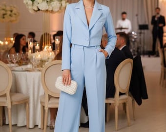 Puede incluir: Un traje azul claro con una chaqueta corta y pantalones de pierna ancha. El traje se usa con un clutch blanco. El fondo muestra un evento formal con mesas, sillas y una banda.