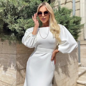 May include: A white dress with long, puffed sleeves and a fitted skirt. The dress has a square neckline and a silver chain necklace. The model is wearing sunglasses.