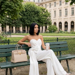 May include: A woman in a white strapless jumpsuit with a large bow detail sits on a green bench, holding a coffee cup. A beige handbag with gold hardware rests beside her. The background features a park setting with trees and a building.
