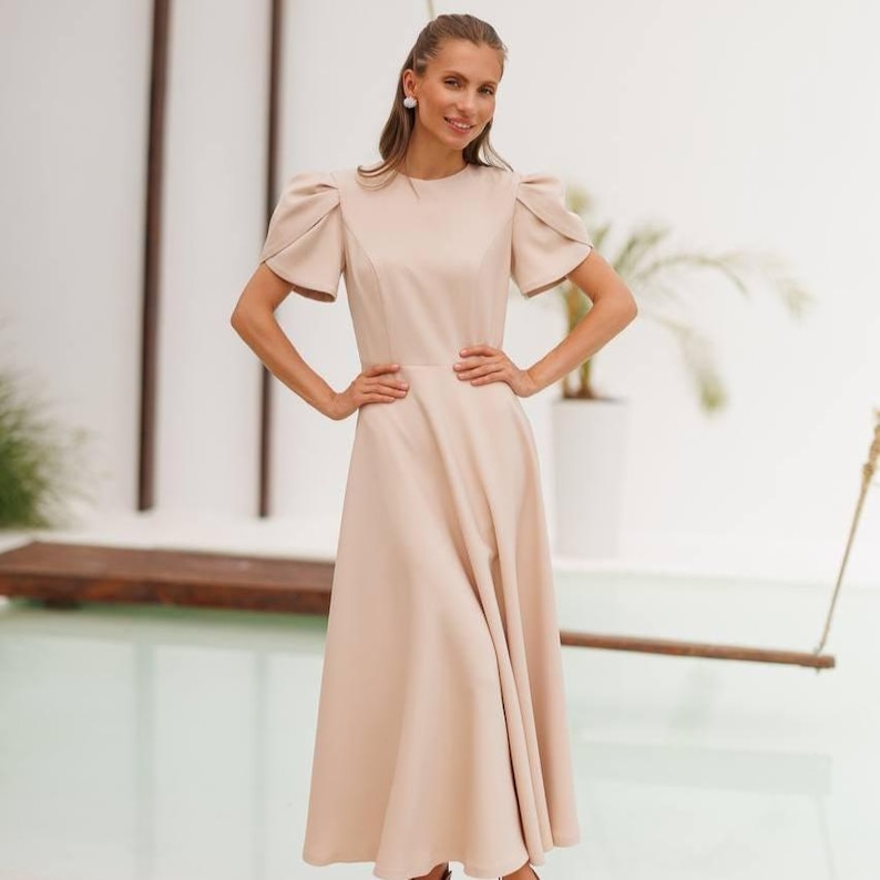 K&ouml;nnte beinhalten: Beiges Midikleid mit kurzen Puff&auml;rmeln und rundem Ausschnitt. Das Kleid hat ein figurbetontes Oberteil und einen ausgestellten Rock. Die Person tr&auml;gt das Kleid und hat die H&auml;nde auf den H&uuml;ften. Der Hintergrund ist eine helle Wand mit einer Pflanze.