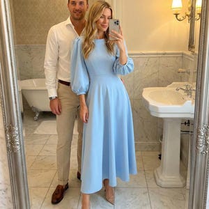 May include: A light blue midi dress with elbow-length sleeves and a fitted waist. The dress is paired with nude heels. A man in a white shirt and tan pants stands behind the woman. The photo is taken in a bathroom with a large mirror.