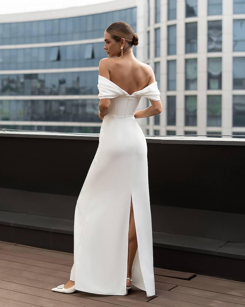 K&ouml;nnte beinhalten: Ein wei&szlig;es schulterfreies Kleid mit hohem Schlitz. Das Kleid hat ein eng anliegendes Oberteil und einen flie&szlig;enden Rock. Das Model steht auf einem Holzdeck mit einem modernen Geb&auml;ude im Hintergrund. Das Kleid wird mit wei&szlig;en Schuhen kombiniert.