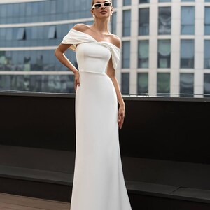 K&ouml;nnte beinhalten: Ein wei&szlig;es schulterfreies Kleid mit einem figurbetonten Oberteil und einem flie&szlig;enden Rock. Das Kleid hat einen drapierten Ausschnitt und ein schlichtes, elegantes Design. Das Model tr&auml;gt Sonnenbrille und Ohrringe.