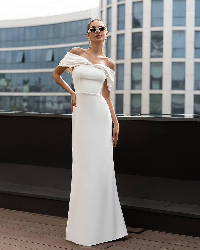 K&ouml;nnte beinhalten: Ein wei&szlig;es schulterfreies Kleid mit einem figurbetonten Oberteil und einem flie&szlig;enden Rock. Das Kleid hat einen drapierten Ausschnitt und ein schlichtes, elegantes Design. Das Model tr&auml;gt Sonnenbrille und Ohrringe.