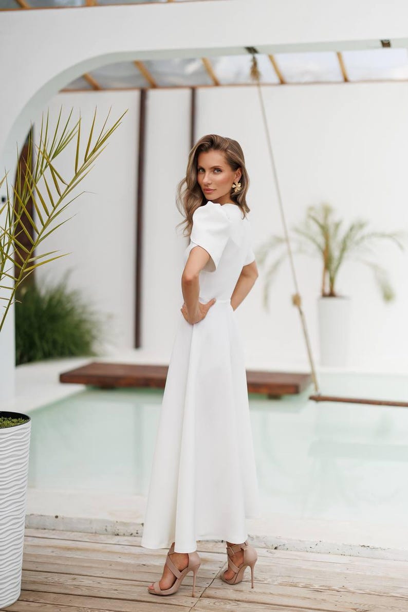 K&ouml;nnte beinhalten: Eine Frau in einem wei&szlig;en Kleid mit kurzen &Auml;rmeln und einem ausgestellten Rock. Das Kleid hat einen V-Ausschnitt und eine taillierte Taille. Sie tr&auml;gt goldene Ohrringe und beige High Heels. Der Hintergrund umfasst einen Pool und tropische Pflanzen.