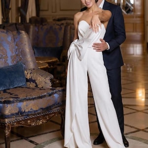 May include: A white strapless jumpsuit with a bow detail at the waist. The wide-leg pants have a split front. A person is wearing the jumpsuit. A person in a navy suit stands behind the person in the jumpsuit.