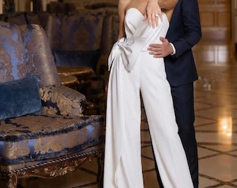 May include: A white strapless jumpsuit with a bow detail at the waist. The wide-leg pants have a split front. A person is wearing the jumpsuit. A person in a navy suit stands behind the person in the jumpsuit.