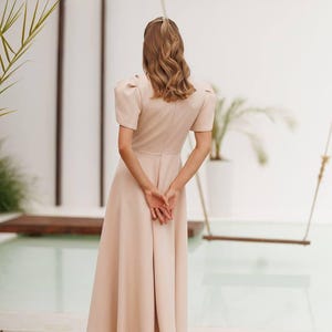 K&ouml;nnte beinhalten: Ein hellbeige Midikleid mit kurzen Puff&auml;rmeln und einem taillierten Oberteil. Das Kleid hat einen ausgestellten Rock und wird mit beigefarbenen High-Heel-Sandalen kombiniert. Das Model steht auf einem Holzdeck, mit einer Schaukel und Gr&uuml;n im Hintergrund.