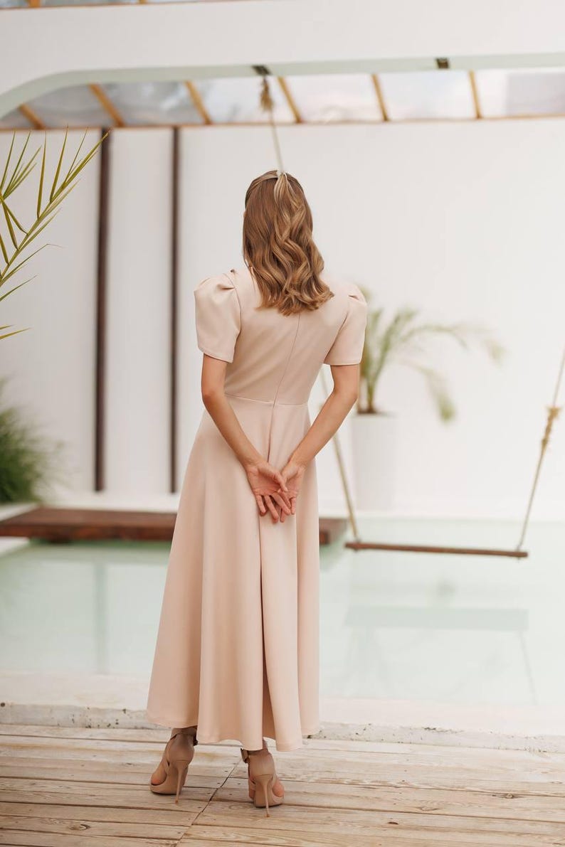 K&ouml;nnte beinhalten: Ein hellbeige Midikleid mit kurzen Puff&auml;rmeln und einem taillierten Oberteil. Das Kleid hat einen ausgestellten Rock und wird mit beigefarbenen High-Heel-Sandalen kombiniert. Das Model steht auf einem Holzdeck, mit einer Schaukel und Gr&uuml;n im Hintergrund.