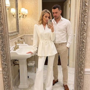 Pode incluir: Um fato branco com um blazer justo e calças de perna larga. O blazer tem decote em V e detalhes de botões. A pessoa está tirando uma selfie com um telefone em um banheiro com um espelho grande.