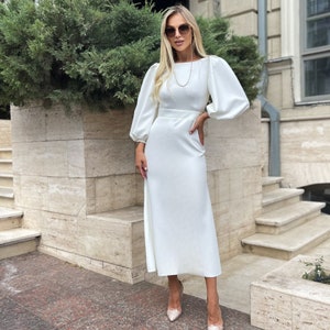 May include: A white midi dress with long sleeves and a boat neckline. The dress has a fitted bodice and a flowing skirt. The model is wearing the dress with a pair of white heels.