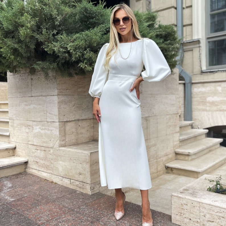 May include: A white midi dress with long sleeves and a boat neckline. The dress has a fitted bodice and a flowing skirt. The model is wearing the dress with a pair of white heels.