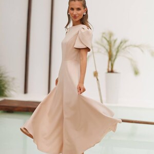 K&ouml;nnte beinhalten: Hellbeige Midikleid mit kurzen Puff&auml;rmeln und flie&szlig;endem Rock. Das Kleid wird mit beigefarbenen High-Heel-Sandalen kombiniert. Der Hintergrund zeigt eine wei&szlig;e Wand, einen Pool und eine Topfpflanze.
