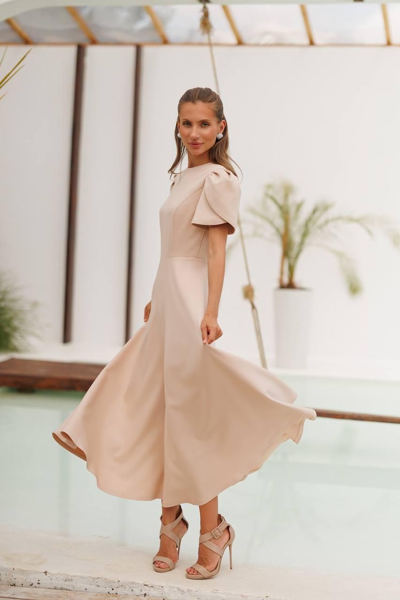 K&ouml;nnte beinhalten: Hellbeige Midikleid mit kurzen Puff&auml;rmeln und flie&szlig;endem Rock. Das Kleid wird mit beigefarbenen High-Heel-Sandalen kombiniert. Der Hintergrund zeigt eine wei&szlig;e Wand, einen Pool und eine Topfpflanze.
