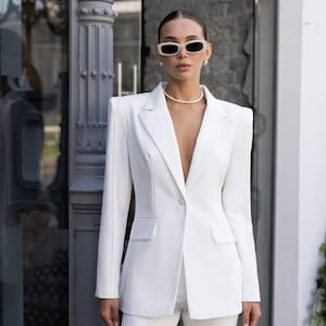 Puede incluir: Una americana blanca entallada con hombros estructurados, cierre de un solo bot&oacute;n, solapas de muesca y dos bolsillos delanteros con solapa. La prenda se combina con pantalones blancos a juego, un collar de perlas y gafas de sol color crema.