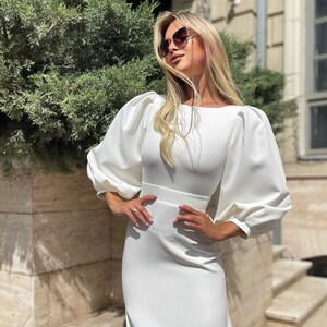 May include: A white dress with long sleeves and a fitted bodice. The sleeves have a puffed design. The dress has a simple, elegant style.