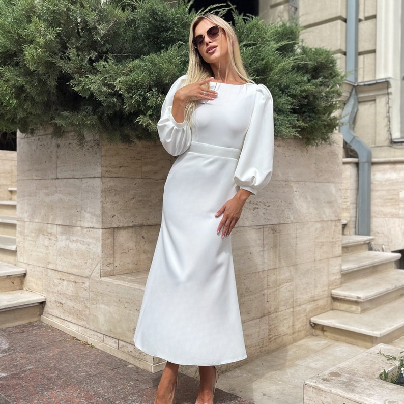 May include: A white midi dress with long sleeves and a fitted bodice. The dress has a slightly flared skirt and a round neckline.