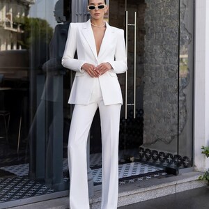 Puede incluir: Un traje blanco entallado compuesto por una americana de un solo bot&oacute;n con hombros estructurados y pantalones a juego de talle alto y pernera ancha. El conjunto se completa con un collar de perlas y gafas de sol rectangulares.