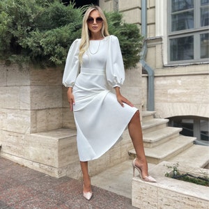 May include: A white midi dress with a fitted bodice and a flowing skirt. The dress has a round neckline and long sleeves with a puffed design. The model is wearing the dress with a pair of nude heels.