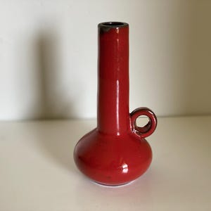 Può includere: Un vaso in ceramica rosso lucido con base bulbosa, lungo collo cilindrico e piccolo manico. Il vaso ha un bordo marrone scuro e una superficie liscia e riflettente. Il vaso è alto circa 18 cm.